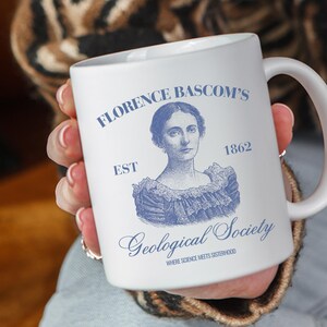 May include: White ceramic mug featuring a blue illustration of Florence Bascom and the text "Florence Bascom's Geological Society" with the year 1862. The mug has a curved handle and is held by a person.