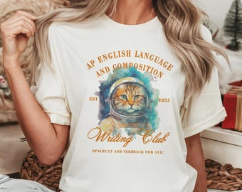 May include: A white t-shirt with a watercolor illustration of a cat wearing an astronaut helmet. The text "AP English Language and Composition Est 1955 Writing Club Spacecat and Feedback for All!" is printed on the shirt.