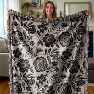 May include: A woven throw blanket featuring a black floral pattern on a light beige background. The design includes large flowers, leaves, and smaller floral accents. The blanket has a fringed edge with multiple colors.