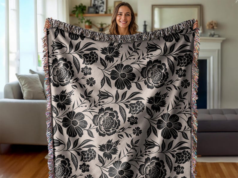 May include: A woven throw blanket featuring a black floral pattern on a light beige background. The design includes large flowers, leaves, and smaller floral accents. The blanket has a fringed edge with multiple colors.