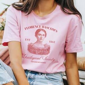 May include: A light pink t-shirt featuring a portrait of Florence Bascom and the text "Florence Bascom's Geological Society" in a reddish-brown color. The shirt also includes the year "1862" and the phrase "Where Science Meets Sisterhood".