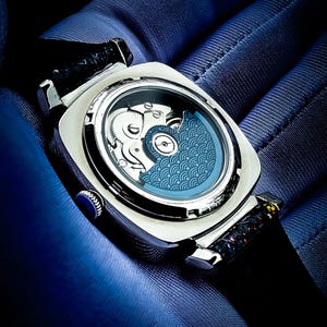 Pode incluir: Um rel&oacute;gio prateado com um mostrador estampado azul e uma pulseira de couro preta. O rel&oacute;gio est&aacute; aberto para revelar o funcionamento interno do mecanismo.