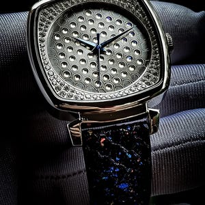 Pode incluir: Um rel&oacute;gio prateado com um mostrador quadrado e uma pulseira de couro preto. O mostrador &eacute; coberto por pequenos diamantes redondos e tem um padr&atilde;o de favo de mel.