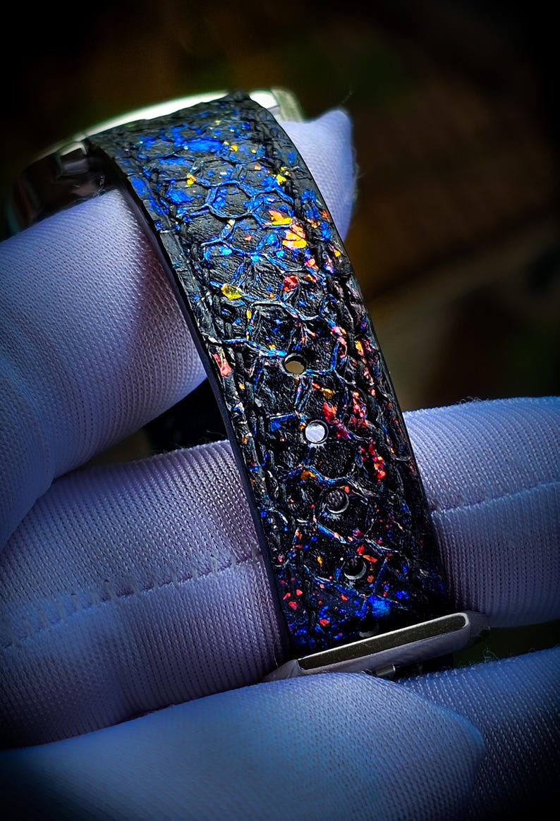 Pode incluir: Uma pulseira de rel&oacute;gio de couro preto com um padr&atilde;o de favo de mel e respingos de tinta iridescente colorida. A pulseira tem dois furos para ajustar o tamanho.