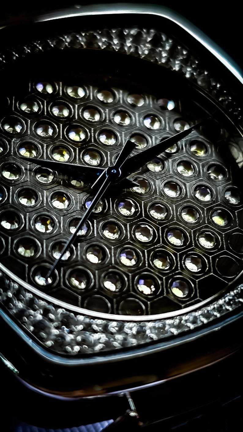 Pode incluir: Um mostrador de rel&oacute;gio preto com um padr&atilde;o hexagonal de pequenos cristais transparentes. Os ponteiros do rel&oacute;gio s&atilde;o pretos e apontam para as 12 horas.