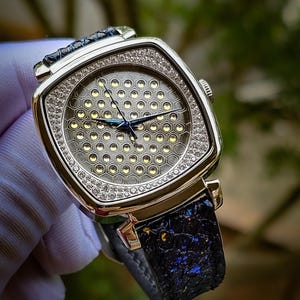 Pode incluir: Um rel&oacute;gio de pulso quadrado prateado com uma pulseira de couro preto. O mostrador do rel&oacute;gio tem um padr&atilde;o de favo de mel com uma moldura de diamantes.