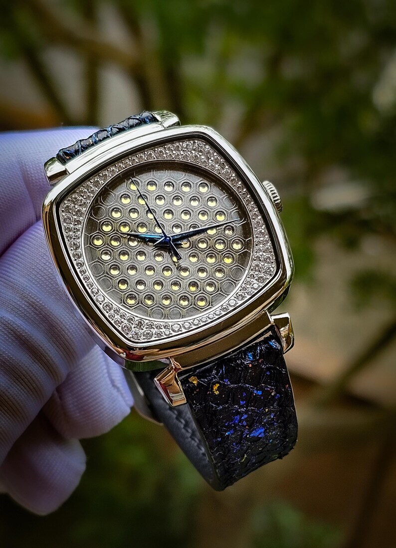 Pode incluir: Um rel&oacute;gio de pulso quadrado prateado com uma pulseira de couro preto. O mostrador do rel&oacute;gio tem um padr&atilde;o de favo de mel com uma moldura de diamantes.