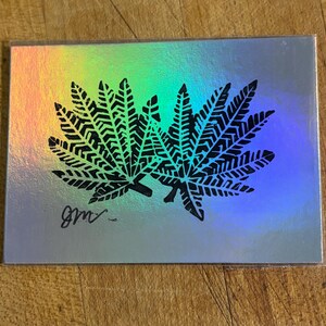 May include: A rectangular card with a reflective holographic surface featuring two black, detailed fern leaf silhouettes printed in the center. The bottom left corner displays a handwritten black signature that reads as the letters GM.