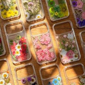 May include: A collection of clear phone cases featuring various arrangements of pressed dried flowers and greenery. Each case displays a unique botanical design, including yellow, pink, red, blue, and green floral patterns embedded in the transparent material.
