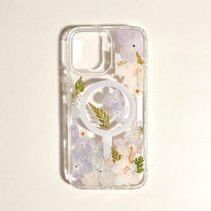 May include: Clear phone case with pressed flowers in shades of white, lavender, and green. The case has a circular design on the back. The case is designed to fit a phone.