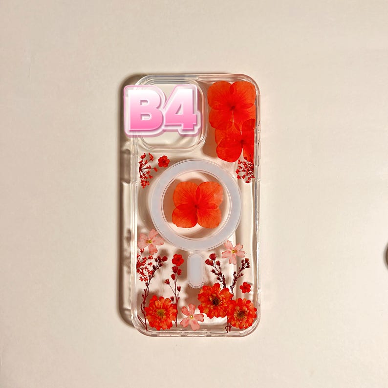 May include: Clear phone case with embedded dried orange flowers and a white circular design. The case has a pink and white text that reads "B4". The case is decorated with small red flowers.