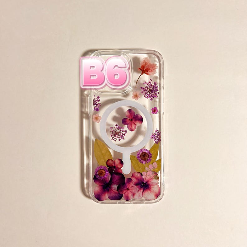 May include: Clear phone case with embedded dried flowers in shades of pink, purple, and yellow. The case has a pink "B6" sticker on the top left. The case has a white circle in the center.