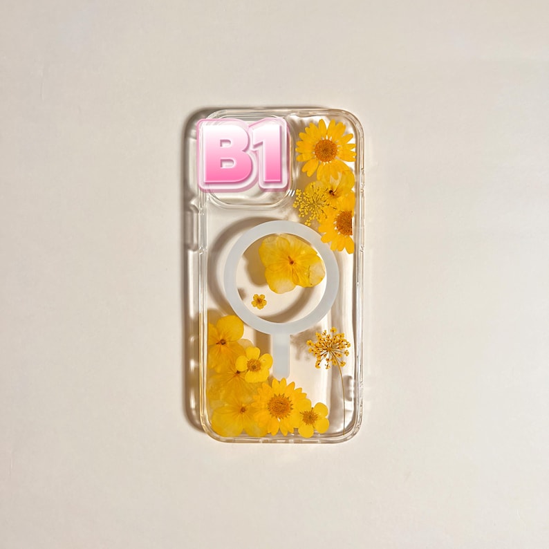 May include: Clear phone case with yellow pressed flowers and the pink text "B1". The case has a circular design and a cluster of yellow flowers. The case is designed to fit a phone.