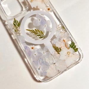 May include: Clear phone case with a floral design. The case features pressed white and purple flowers, along with green leaves. A white circular MagSafe ring is visible in the center of the case.