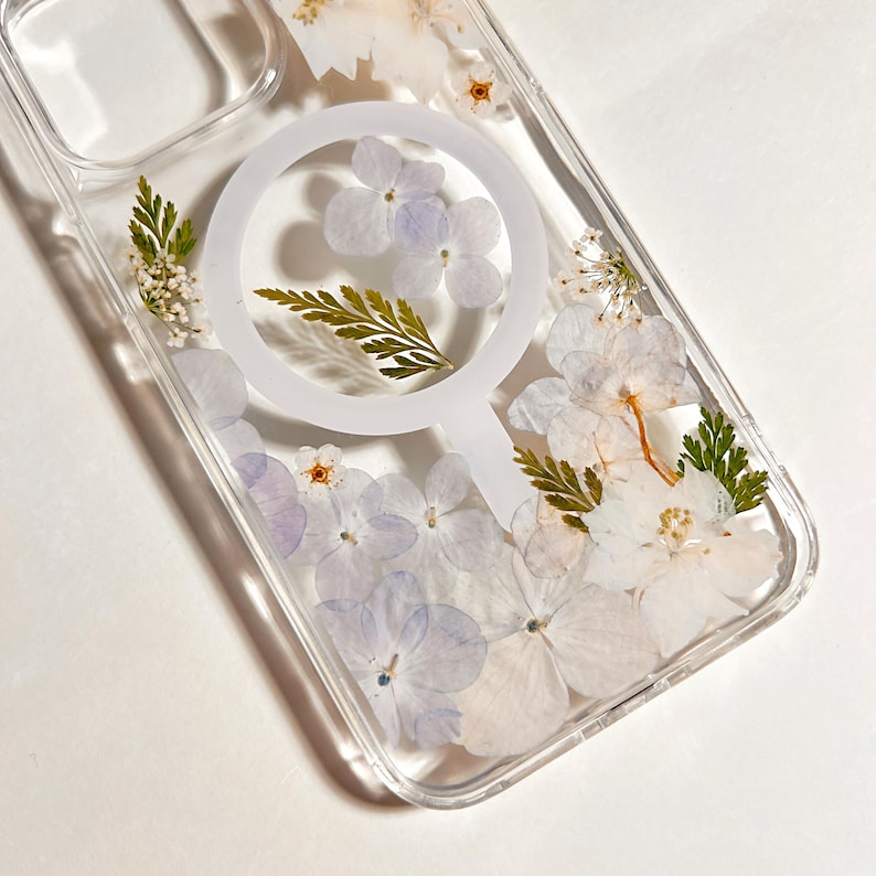 May include: Clear phone case with a floral design. The case features pressed white and purple flowers, along with green leaves. A white circular MagSafe ring is visible in the center of the case.