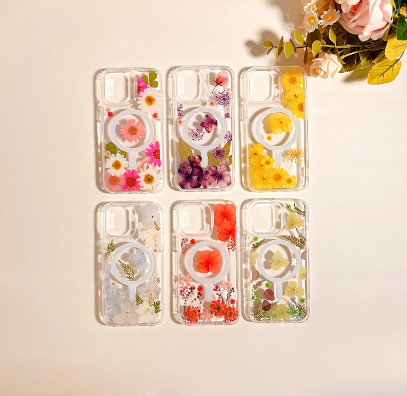 May include: Clear phone cases with embedded dried flowers in various colors, including pink, purple, yellow, and red. Each case features a circular design element. The cases are arranged in a grid, showcasing the floral arrangements.