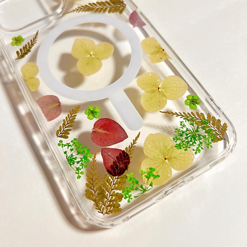 May include: Clear phone case with a white circular design and embedded dried flowers. The case features yellow, green, and red botanical elements, creating a natural and artistic aesthetic. The case is designed to protect the phone while showcasing the floral arrangement.