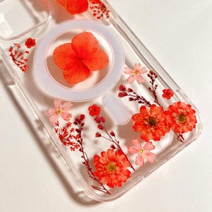 May include: Clear phone case with embedded dried orange flowers and accents. The case features a white circular MagSafe ring and a rectangular button area. The flowers are arranged in a decorative pattern, adding a natural and artistic touch to the phone accessory.