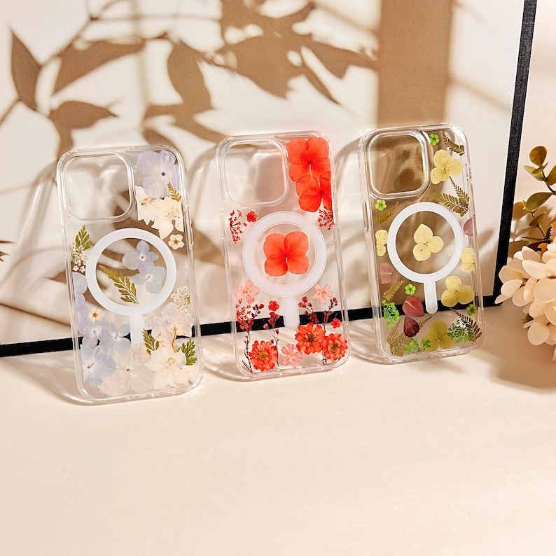 May include: Three clear phone cases with embedded dried flowers in shades of red, yellow, and purple. Each case features a white circular design. The cases are displayed on a light-colored surface.