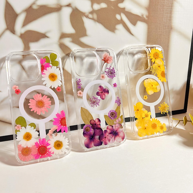 May include: Three clear phone cases with embedded dried flowers. The cases feature a white circular design. One case has pink and white daisies, another has purple flowers, and the third has yellow flowers.