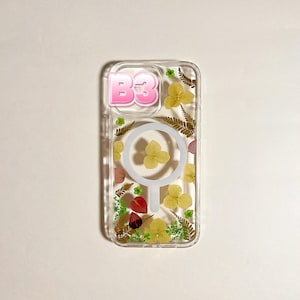 May include: Clear phone case with dried flowers and greenery. The case has a pink "B3" graphic and a white circular design. Yellow and red flowers are visible within the case.