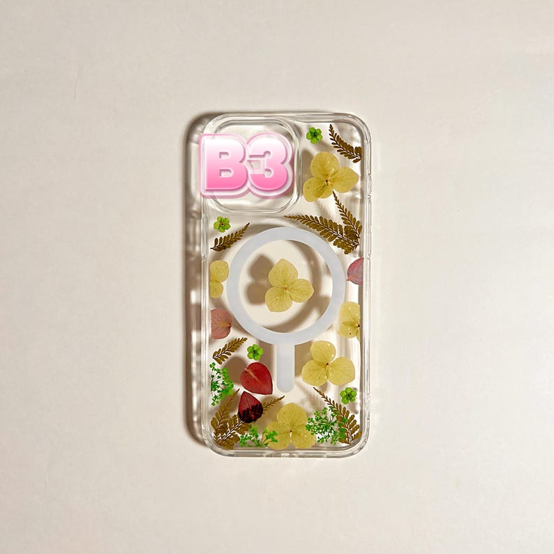 May include: Clear phone case with dried flowers and greenery. The case has a pink "B3" graphic and a white circular design. Yellow and red flowers are visible within the case.