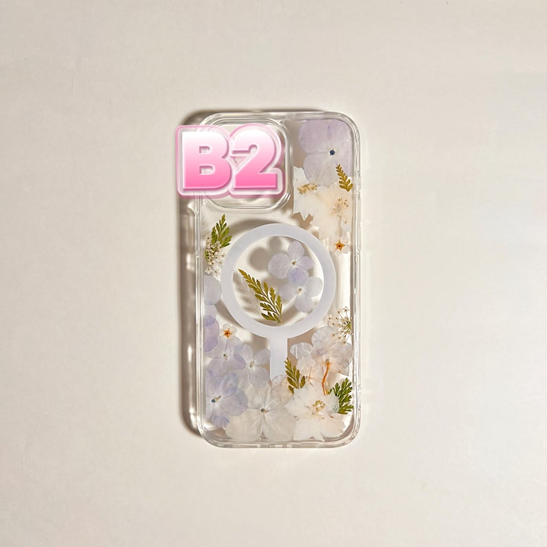 May include: Clear phone case with a floral design. The case features pressed white and purple flowers, green leaves, and a white circular MagSafe charger. A pink sticker with the text "B2" is on the top left corner.