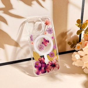 May include: A clear phone case adorned with pressed flowers in shades of pink and purple. The case features a circular design with a white border and a collection of dried floral elements, including petals and leaves.