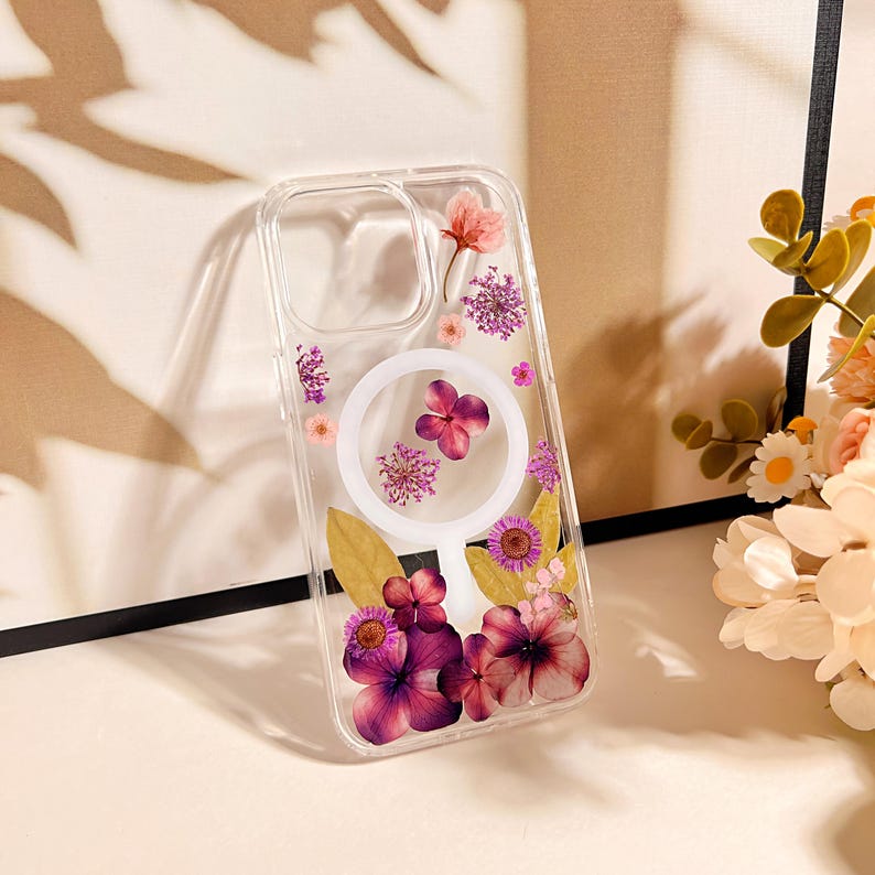 May include: A clear phone case adorned with pressed flowers in shades of pink and purple. The case features a circular design with a white border and a collection of dried floral elements, including petals and leaves.