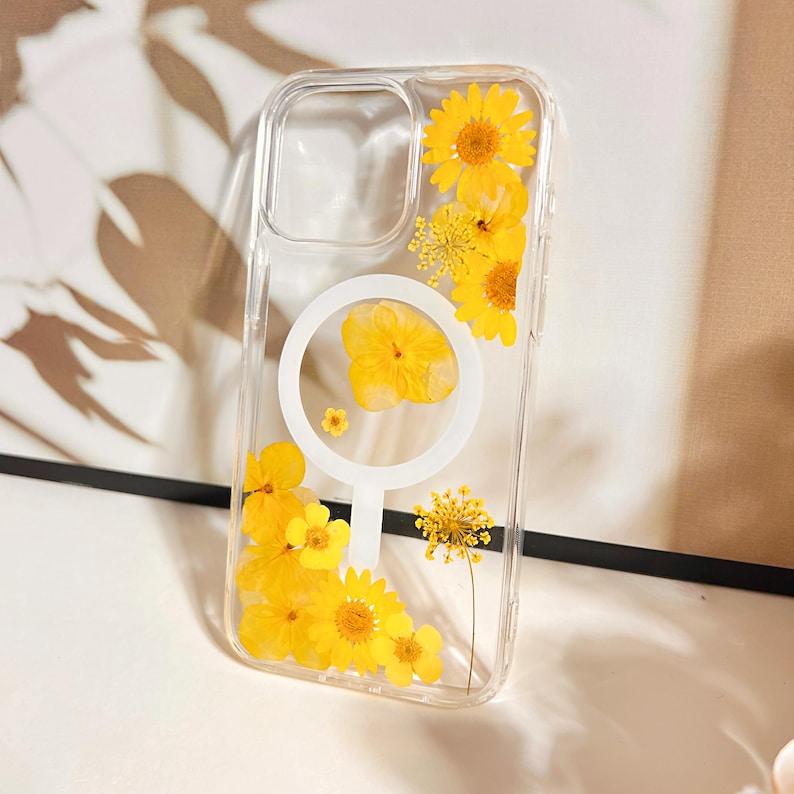 May include: Clear phone case with embedded yellow dried flowers. The case features a white circular design and a clear border. The flowers include daisies and other small yellow blooms, creating a natural, floral aesthetic.