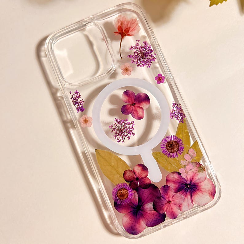 May include: Clear phone case with embedded dried flowers in shades of pink, purple, and yellow. The case features a white circular design. The flowers are arranged throughout the case, creating a decorative and natural aesthetic.
