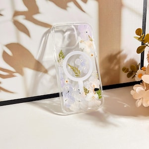 May include: Clear phone case with a floral design. The case features pressed white and purple flowers and green leaves. A white circular magnetic charging ring is visible on the back of the case.