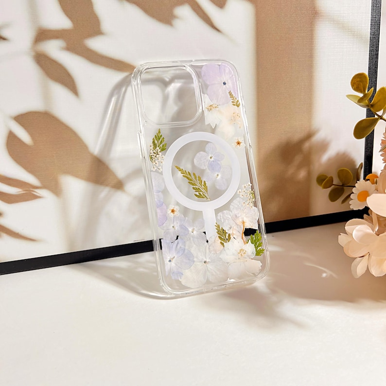 May include: Clear phone case with a floral design. The case features pressed white and purple flowers and green leaves. A white circular magnetic charging ring is visible on the back of the case.