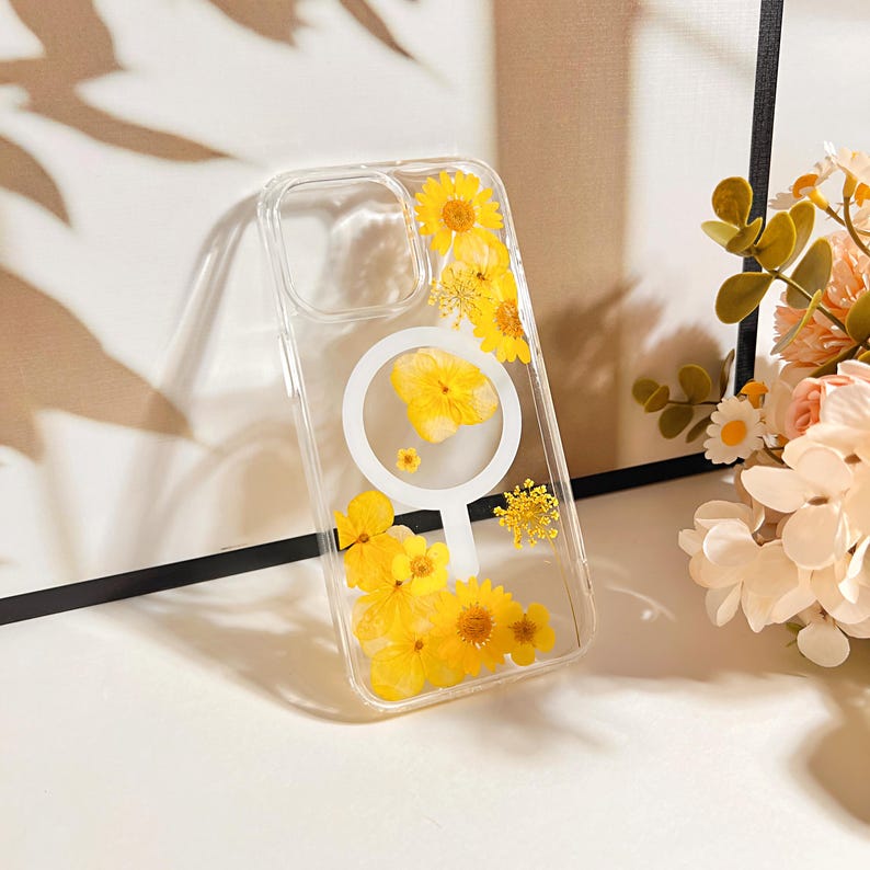May include: Clear phone case with yellow dried flowers embedded. The case has a white circular design on the back. The flowers are various sizes and shapes, adding a natural and artistic touch to the phone accessory.
