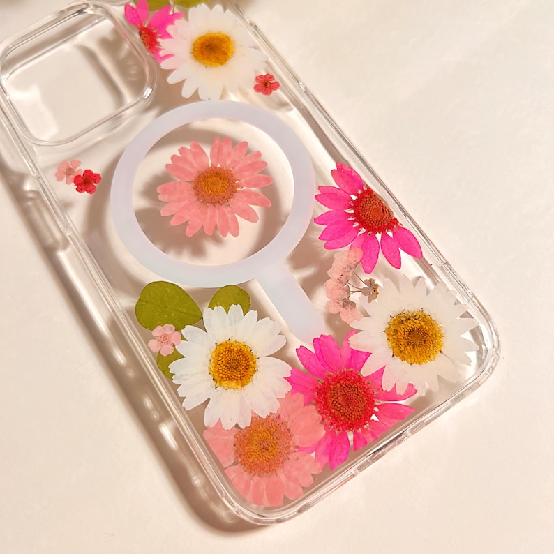 May include: Clear phone case decorated with pressed pink and white daisies, small red flowers, and green leaves. The case has a white circular design in the center.
