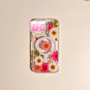 May include: Clear phone case with a floral design. The case features a pink and white daisy arrangement with the text "B5" in a pink bubble letter font. The case has a circular design with a pink flower in the center.