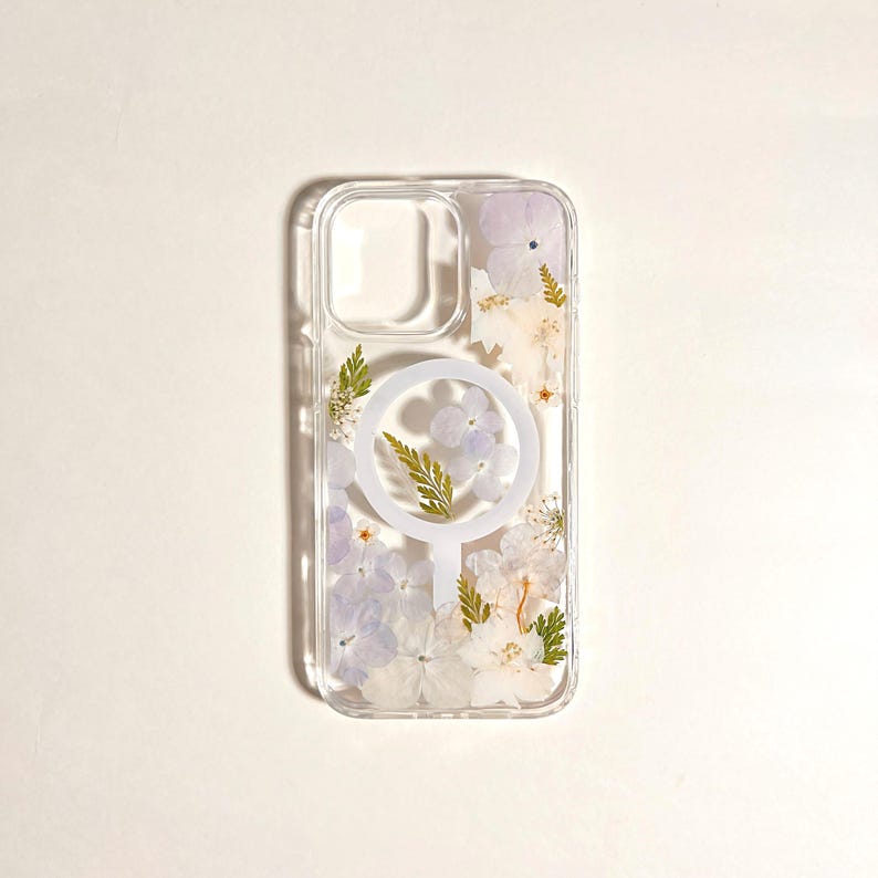 May include: Clear phone case with pressed flowers in shades of white, lavender, and green. The case has a circular design on the back. The case is designed to fit a phone.