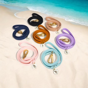 Può includere: Sei guinzagli per cani in silicone colorati con accessori dorati e un logo dorato. I guinzagli sono disposti in cerchio su una spiaggia di sabbia bianca.