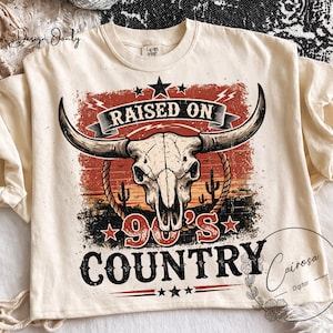 May include: Cream-colored t-shirt with a graphic featuring a bull skull, cactus, and the text "RAISED ON 90'S COUNTRY." The design has a vintage, distressed look with red, orange, and black accents. The shirt has a crew neck.