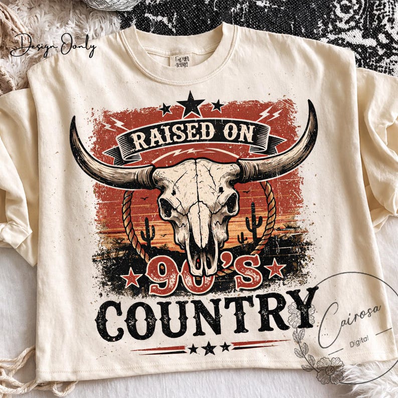 May include: Cream-colored t-shirt with a graphic featuring a bull skull, cactus, and the text "RAISED ON 90'S COUNTRY." The design has a vintage, distressed look with red, orange, and black accents. The shirt has a crew neck.