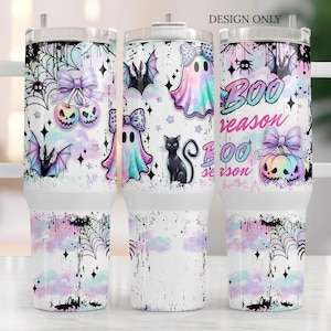 May include: A stainless steel tumbler with a handle featuring a pastel Halloween pattern of ghosts, bats, a black cat, spiderwebs, and pumpkins with bows. The text reads BOO season in pink and blue gradient lettering against a white background.