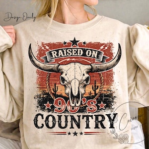 May include: Beige sweatshirt featuring a graphic design with a bull skull, cacti, and the text "RAISED ON 90'S COUNTRY." The design incorporates red, black, and white elements, with stars and a banner. The sweatshirt has long sleeves.