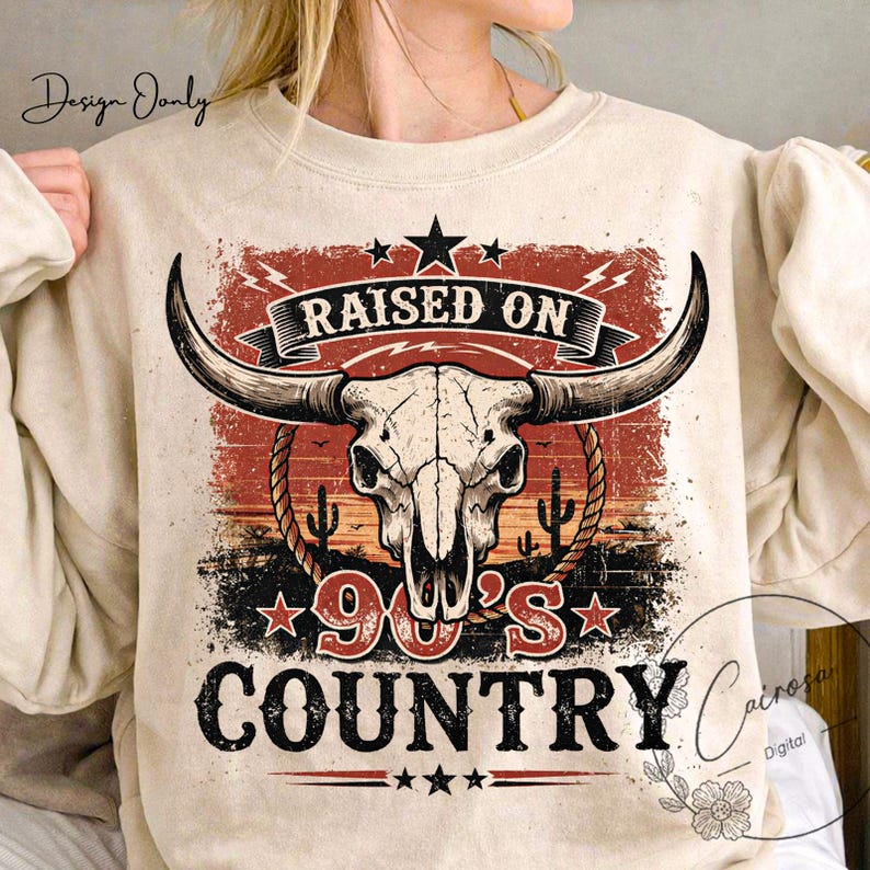 May include: Beige sweatshirt featuring a graphic design with a bull skull, cacti, and the text "RAISED ON 90'S COUNTRY." The design incorporates red, black, and white elements, with stars and a banner. The sweatshirt has long sleeves.