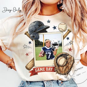May include: Cream-colored t-shirt featuring a baseball-themed design. The design includes a baseball helmet, baseball bat, baseball, and a baseball glove. A photo of a football player is in the center. The words "GAME DAY" are on a banner.