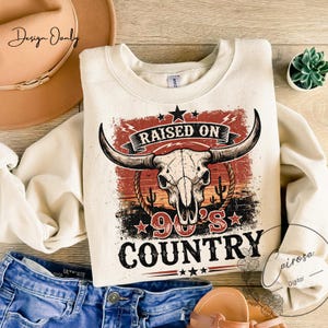 May include: Cream-colored sweatshirt with a graphic featuring a bull skull, stars, and the text "RAISED ON 90'S COUNTRY." The design includes a red and orange sunset-like background with cacti. The sweatshirt is paired with blue denim jeans and brown sandals.