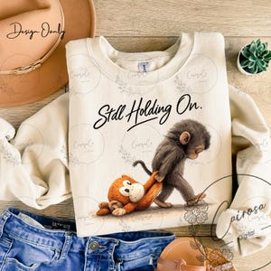May include: Cream-colored sweatshirt with the text "Still Holding On." features a cartoon monkey pulling another monkey. The design includes floral accents. The sweatshirt is paired with blue jeans and a brown hat.
