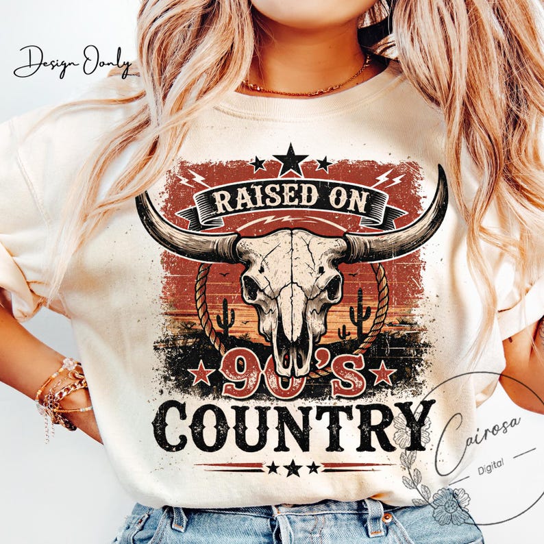 May include: Cream-colored t-shirt featuring a graphic design with a bull skull, cacti, and the text "RAISED ON 90'S COUNTRY." The design incorporates red, black, and brown tones, with stars and lightning bolts. The shirt has a crew neck.