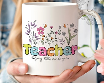 May include: White ceramic mug with the word "Teacher" in colorful letters, surrounded by floral and butterfly illustrations. The phrase "helping little minds grow" is below the word "Teacher".