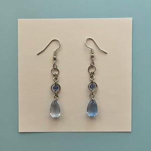 May include: A pair of silver-coloured drop earrings. Each earring has a hook, a series of small rings, a blue bead, and a teardrop-shaped blue glass pendant. The earrings are presented on a white card against a blue background.
