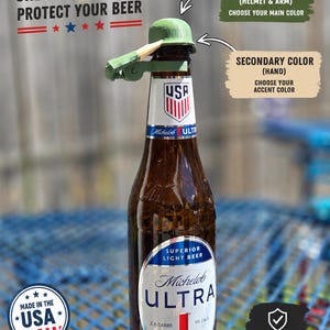 May include: A green plastic bottle topper shaped like a military helmet with an attached arm, placed on a Michelob Ultra beer bottle. Text on the image reads: TWO COLORS ONE MISSION: PROTECT YOUR BEER, PRIMARY COLOUR, SECONDARY COLOUR, MADE IN THE USA, KEEPS BUGS & DEBRIS OUT OF YOUR DRINK.